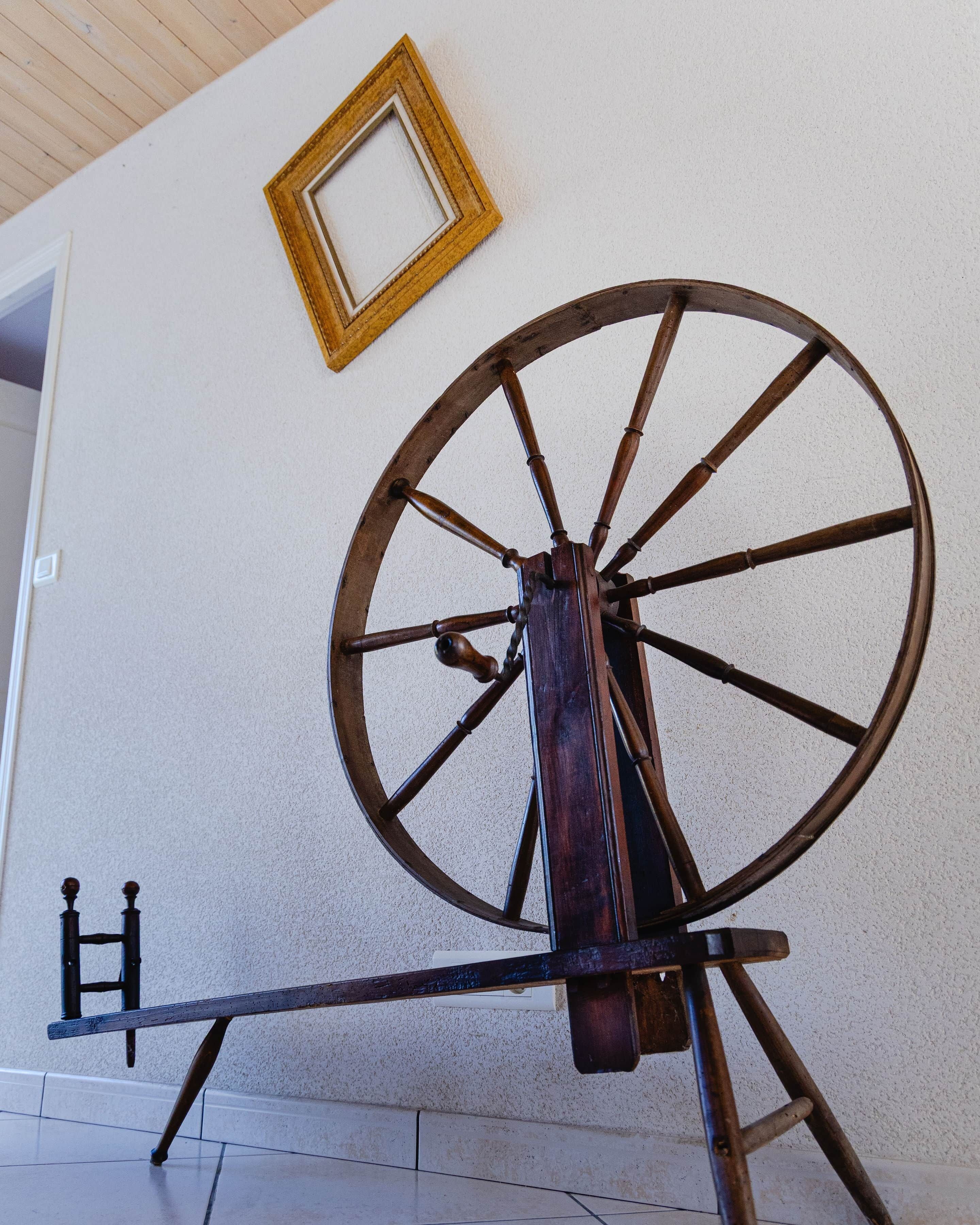 Een groot houten wielconstructie, mogelijk een decoratief object of onderdeel van een mechanische installatie, bevestigd aan een standaard voor een witte muur, met daarboven een lijst met een afbeelding.