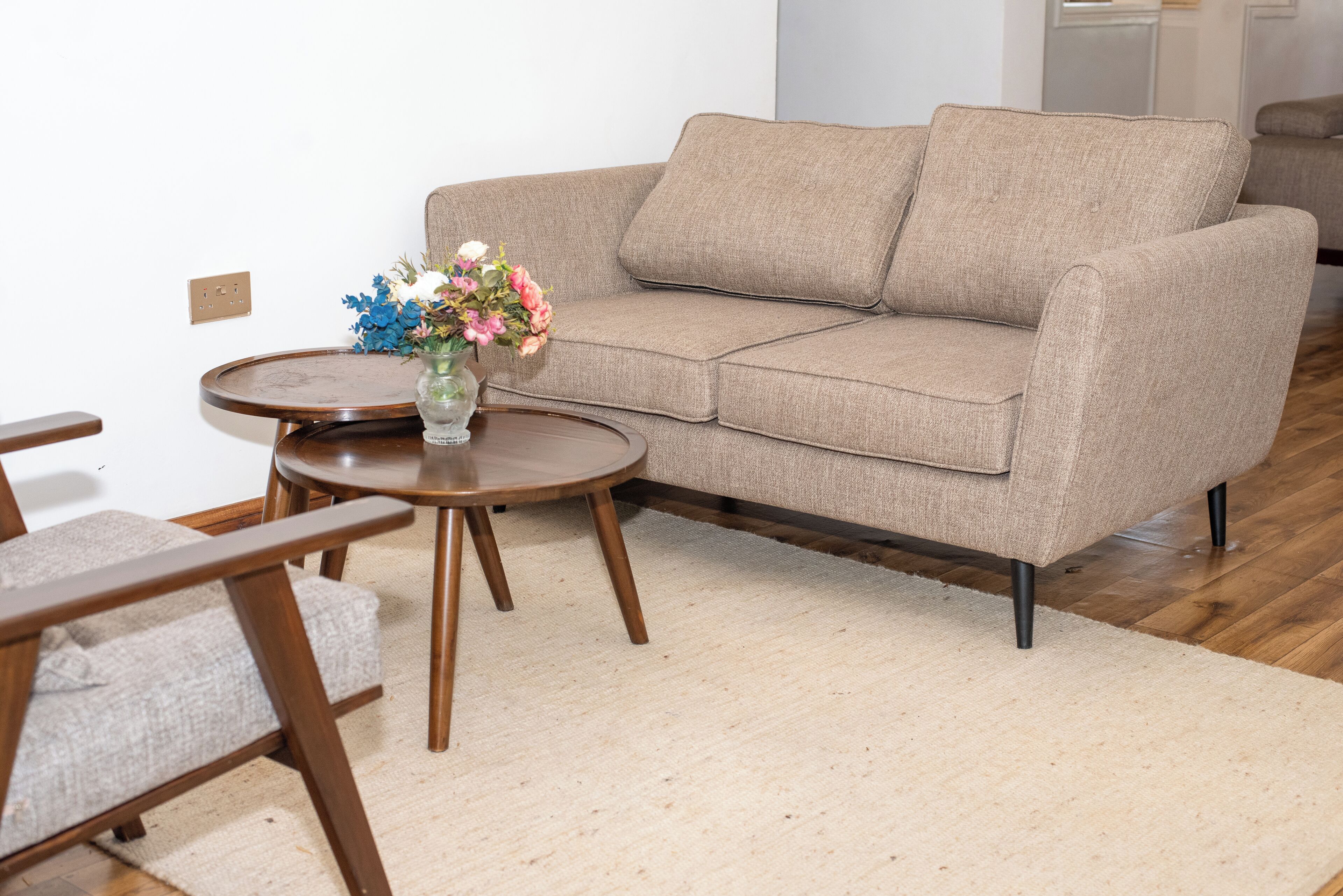 A beige sofa with wooden legs, a round wooden table with a vase of flowers, and a light-colored rug on a wooden floor.