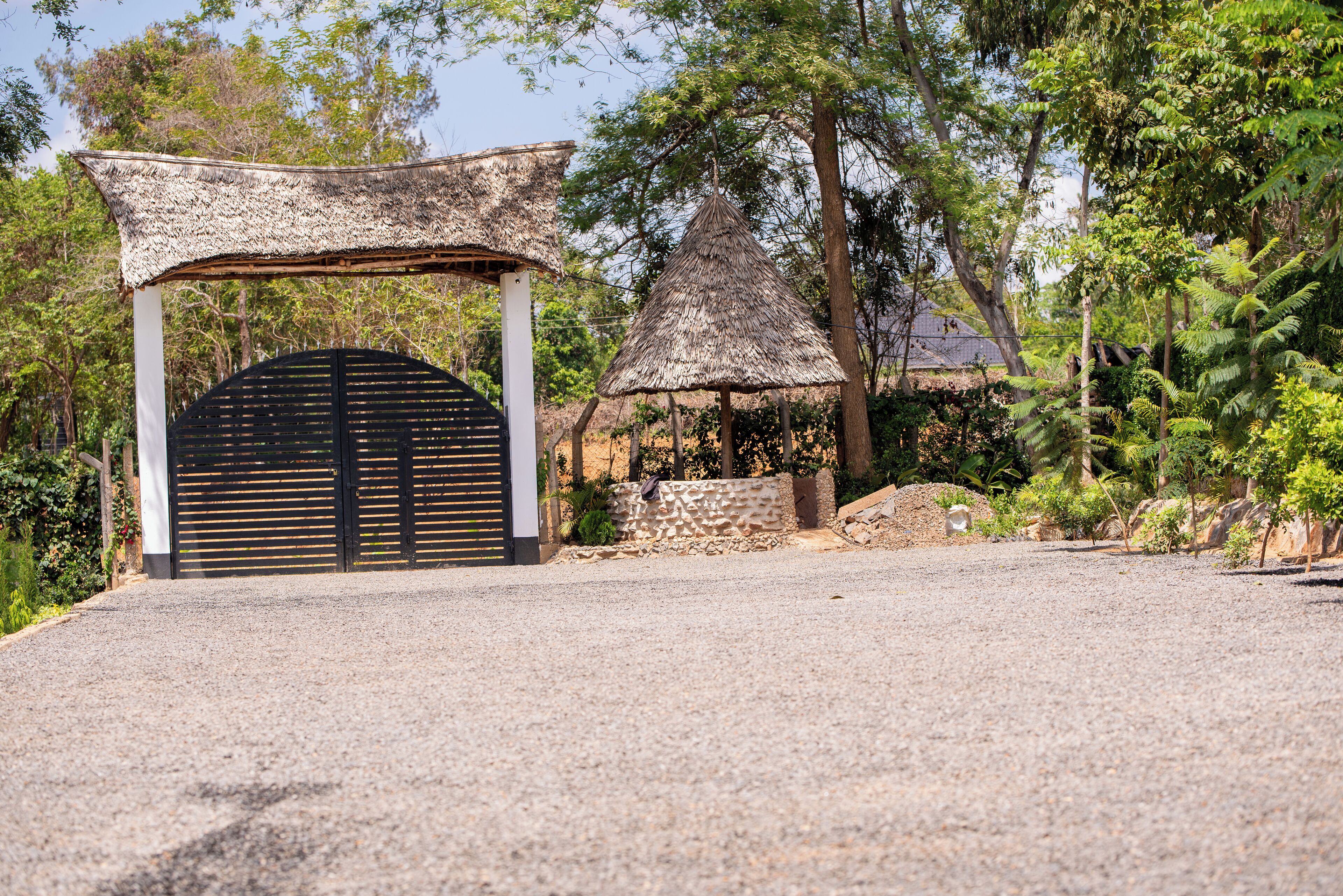 A gravel driveway leading to a building with a black gate and a thatched-roof structure.