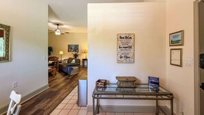 A living room with a sofa, coffee table, and a glass-top table with decorative items. There is a ceiling fan and a framed sign on the wall.