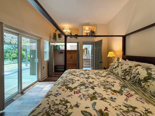 A bedroom with a four-poster bed, a sliding glass door leading to a balcony, and a view of a garden.