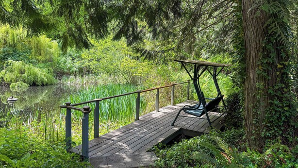 A wooden deck with a swing chair overlooking a pond surrounded by lush greenery.