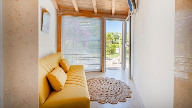 Une pièce avec un canapé jaune, une grande fenêtre avec des stores, un ventilateur de plafond et une vue sur la verdure à l’extérieur.