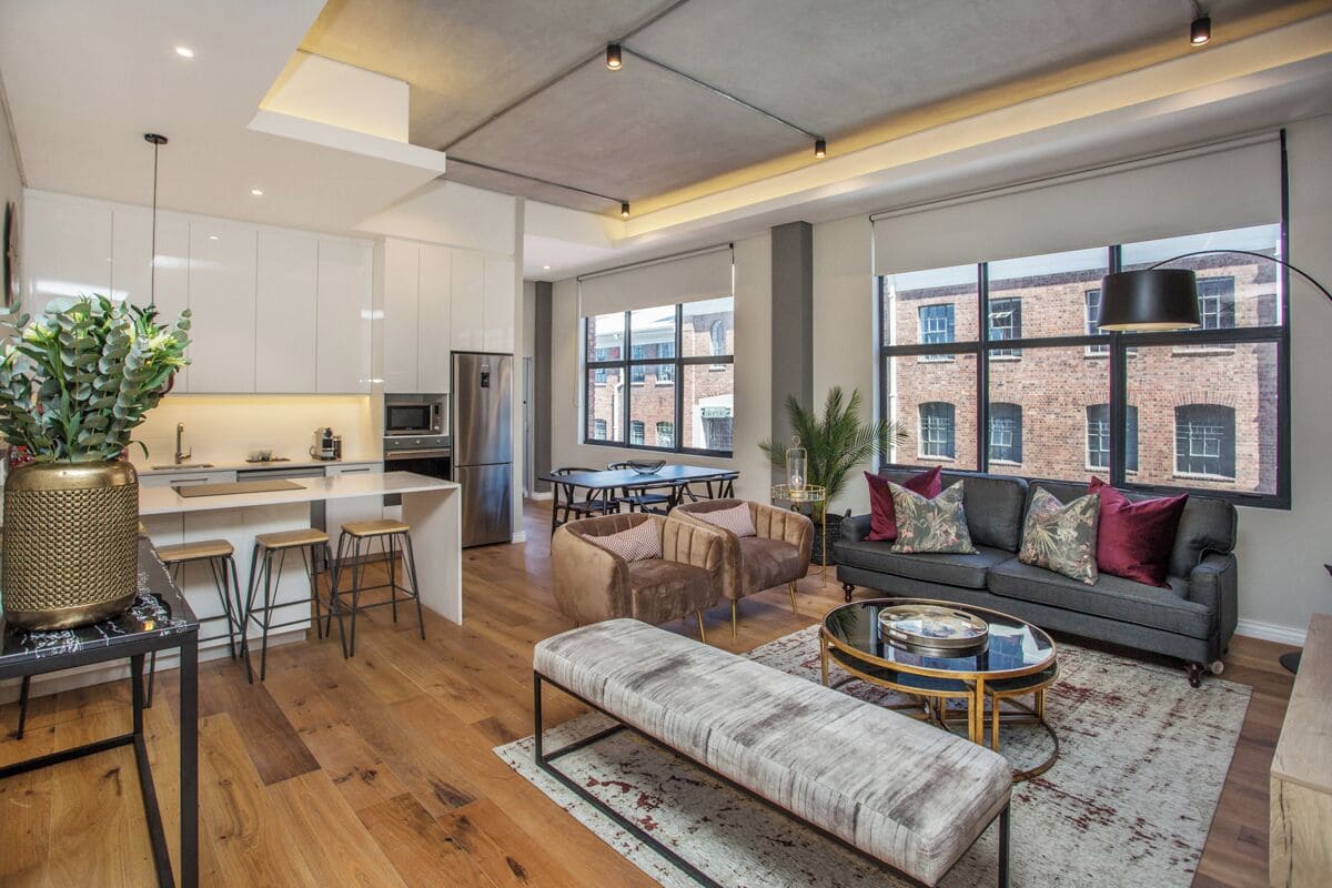 Een modern appartement met keuken, eetruimte en woonkamer. Het heeft een groot raam met uitzicht op de stad, een bank, een salontafel en een lange bank.