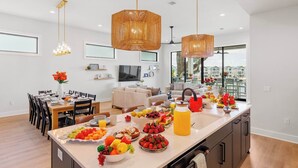 A modern kitchen with a large island, hanging wicker chandeliers, and a dining area with a TV and seating.