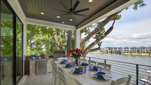 A covered outdoor dining area with a view of a body of water, a ceiling fan, and a stone countertop with kitchen appliances.
