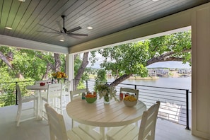 A covered outdoor dining area with white furniture, a table with bowls of fruit and flowers, and a view of a river and buildings.