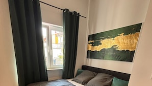 Un dormitorio con cama, mesita de noche, lámpara, ventana con cortinas y un cuadro colgado en la pared.