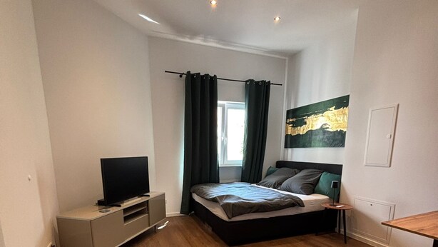 Un dormitorio moderno con cama, televisor sobre un mueble bajo, una mesita y un cuadro en la pared.