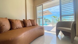 A modern living room with a brown leather sofa and a grey armchair, large sliding glass doors leading to a pool area. - NYC Park Ave Beach Apt •3BR • Private Pool • Oceanview in Ponta Negra Natal (Ponta Negra Natal)