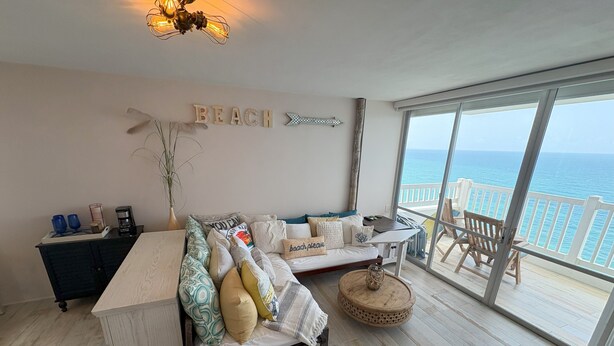 A spacious room with a large window overlooking the sea, a cozy sofa with various cushions, a wooden coffee table, and a small side table.