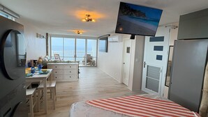 A modern, well-lit room with a bed, a television mounted on the wall, a kitchen area, and a view of the sea through large windows.