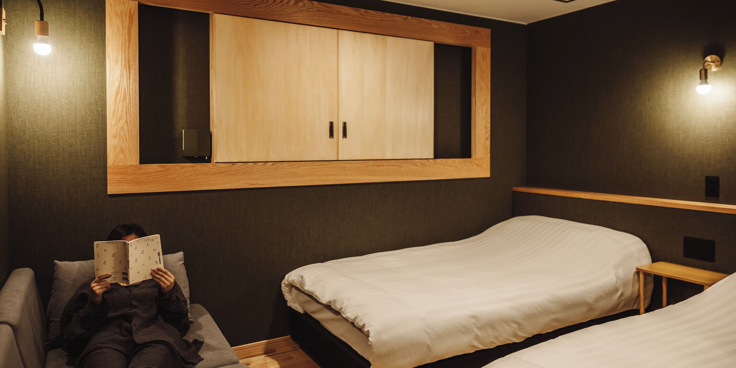 A bedroom with two beds, a wooden wardrobe, and a person reading a book.