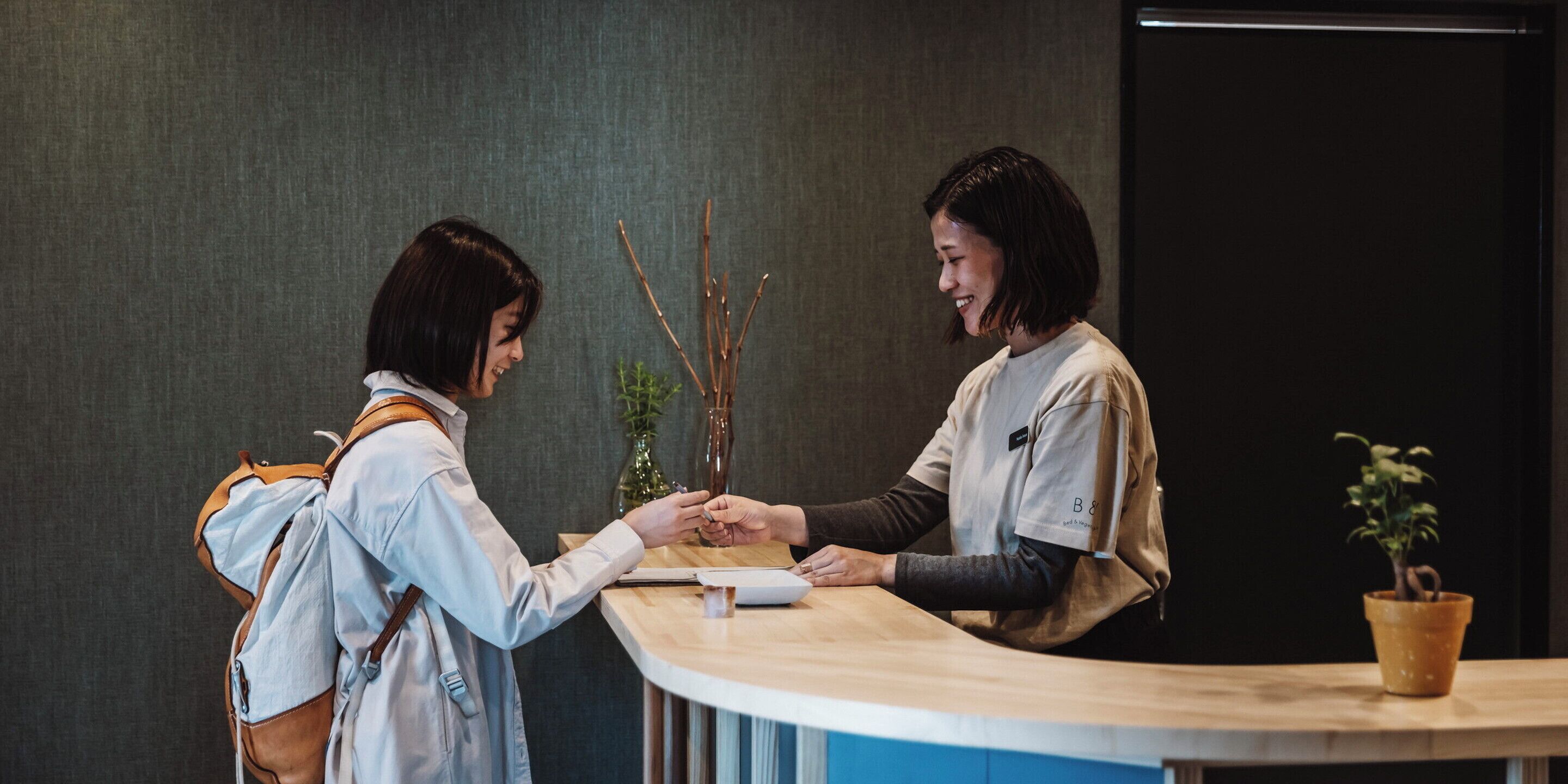 Two individuals at a reception desk with a plant in the background.