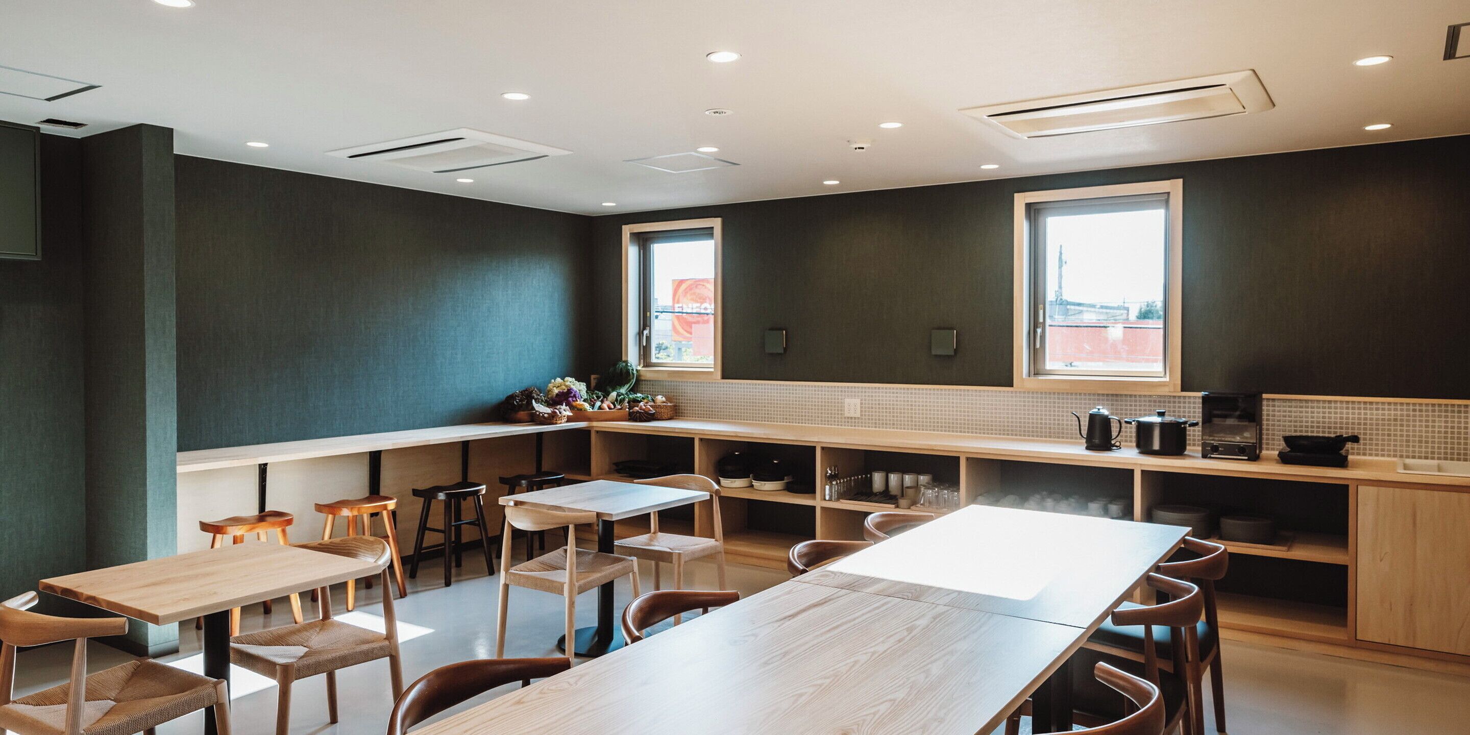 A modern restaurant interior with a long communal table, individual seating, and a kitchen area visible through the counter.