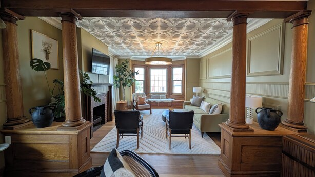 A spacious living room with a fireplace, large windows, and a high ceiling with intricate moldings.