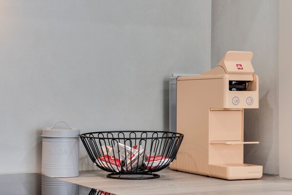 A kitchen area with a coffee machine, a metal bowl with sugar containers, and a trash bin.