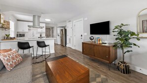 A modern living room with a wooden coffee table, a sofa with patterned cushions, a dining area with bar stools, and a kitchen with white cabinets and stainless steel appliances. - Optimist Abode 4: Chic Stay in the <3 of CLT with Greenway Access (Charlotte)