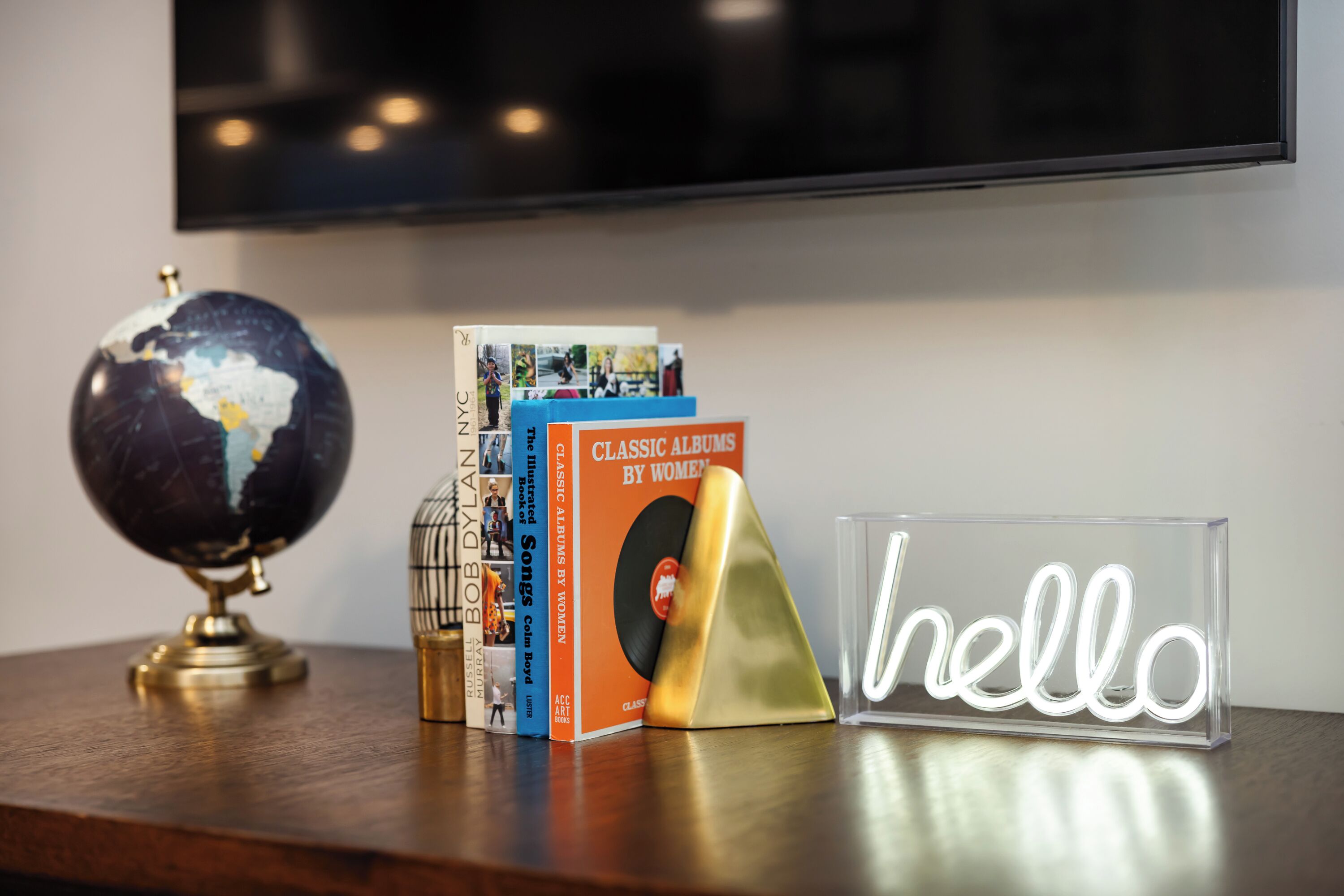 Une étagère en bois sur laquelle on trouve un globe terrestre, une pile de livres intitulée « Albums classiques d'auteures » et un néon affichant le mot « bonjour ».