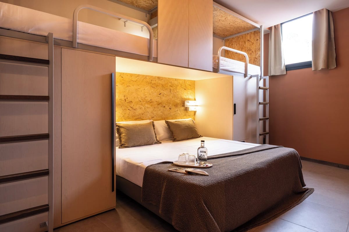 A compact hotel room with bunk beds, a bed with a headboard, a bedside table with a bottle and glasses, and a window with curtains.