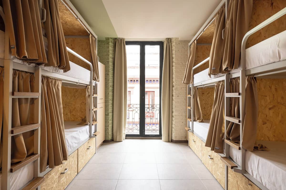 A dormitory room with bunk beds, wooden walls, a tiled floor, and a view of a balcony with metal railings.