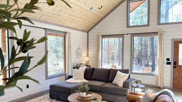 Ein geräumiges Wohnzimmer mit hoher Decke, großen Fenstern mit Blick auf einen Wald, ein graues Modularesoap, ein brauner Ledersessel und ein kleiner runder Couchtisch.