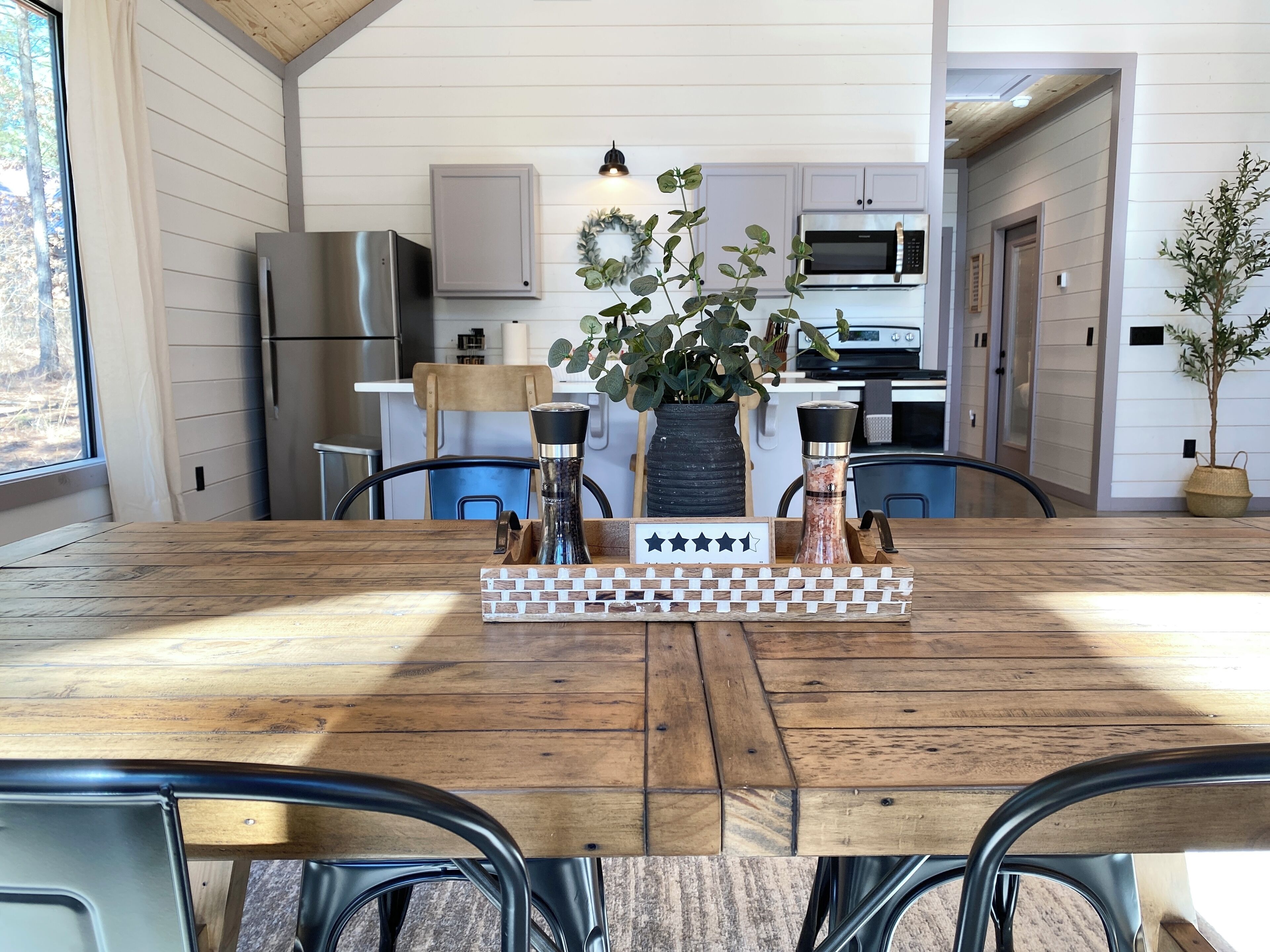 A dining area with a wooden table, metal chairs, a chessboard centerpiece, and a kitchen in the background.