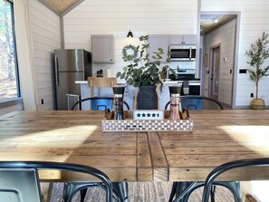 Un coin repas avec une table en bois, des chaises en métal, un jeu d’échecs en guise de décoration centrale et une cuisine en arrière-plan.