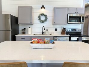Une cuisine moderne avec des meubles gris, un réfrigérateur en acier inoxydable, un four et un four à micro-ondes. On y trouve un îlot central blanc sur lequel est posé un saladier rempli de collations.