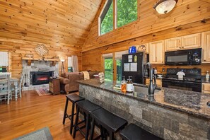 A wooden cabin interior with a stone fireplace, a kitchen with modern appliances, and a bar area with stools. - *Gorgeous 3BD Cabin Near LAKE w/ Pool Access* (Sevierville)