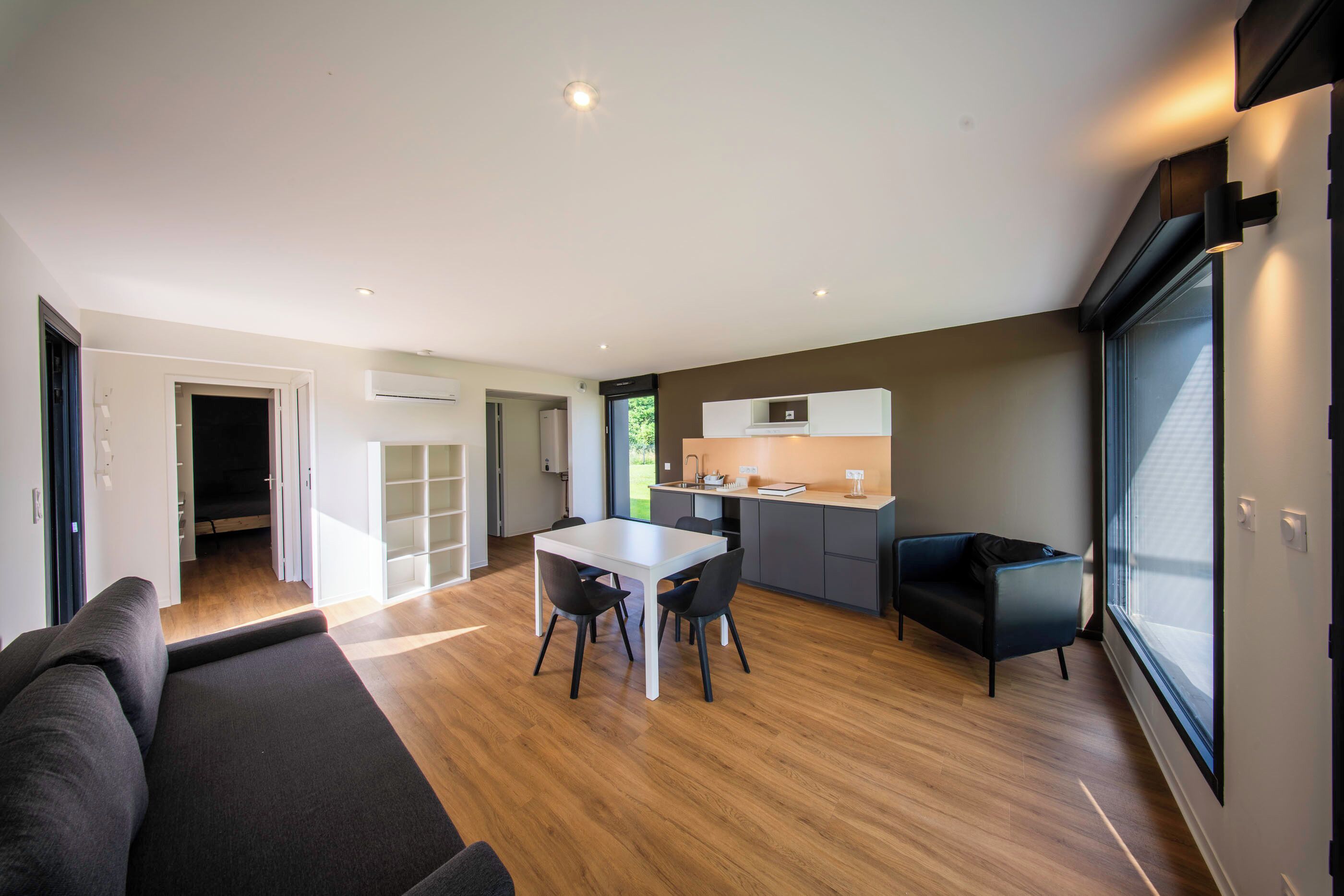 A modern living room with a sofa, a dining table with chairs, a kitchen area with a countertop and cabinets, and a large window providing natural light.