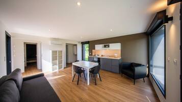 A modern living room with a sofa, a dining table with chairs, a kitchen area with a countertop and cabinets, and a large window providing natural light.