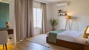 A hotel room with a bed, a desk, a standing lamp, a potted plant, and a window with curtains. - Prime Residence Golf Kattameya (Shurtah al-Dakhlah)