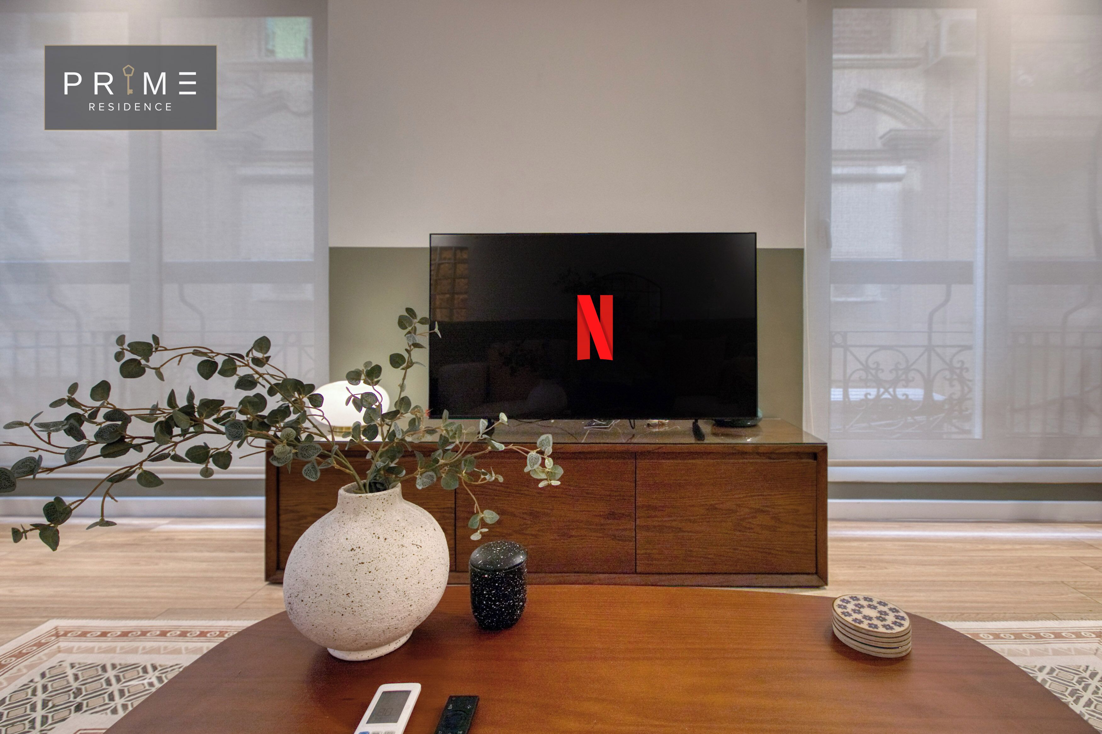 A living room with a television displaying the Netflix logo, a wooden coffee table, a white vase with green foliage, and a small black decorative item.