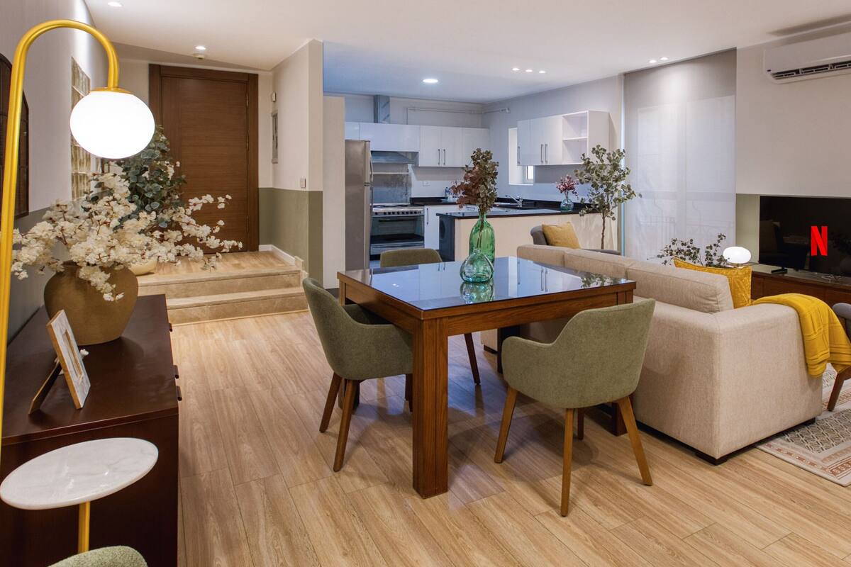 Een moderne woonkamer met een glazen eettafel, groene en beige stoelen, een bank en een televisie.