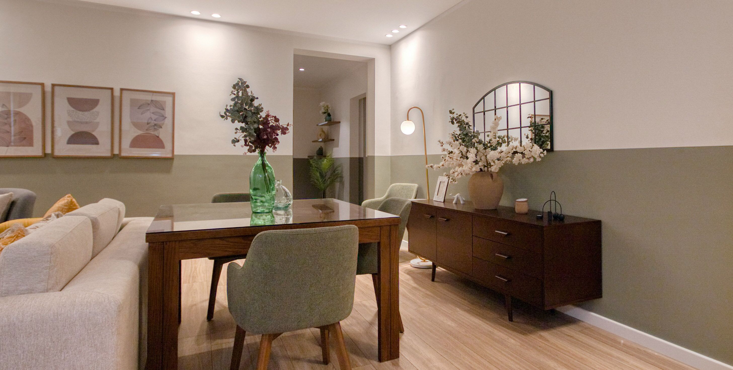 A dining area with a wooden table, chairs, and a sofa. There are framed pictures on the wall and a vase with flowers on the table.