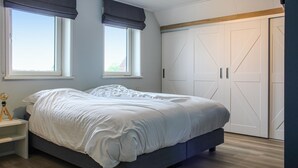 A bedroom with a bed, two windows with dark curtains, a wardrobe with a geometric design, and a wooden floor.