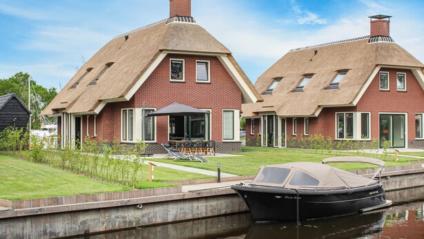 A house with a thatched roof, a boat docked in a canal, and a grassy yard.