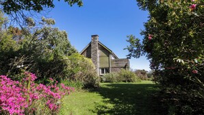 Una casa con chimenea de piedra, rodeada de exuberante vegetación y arbustos en flor.