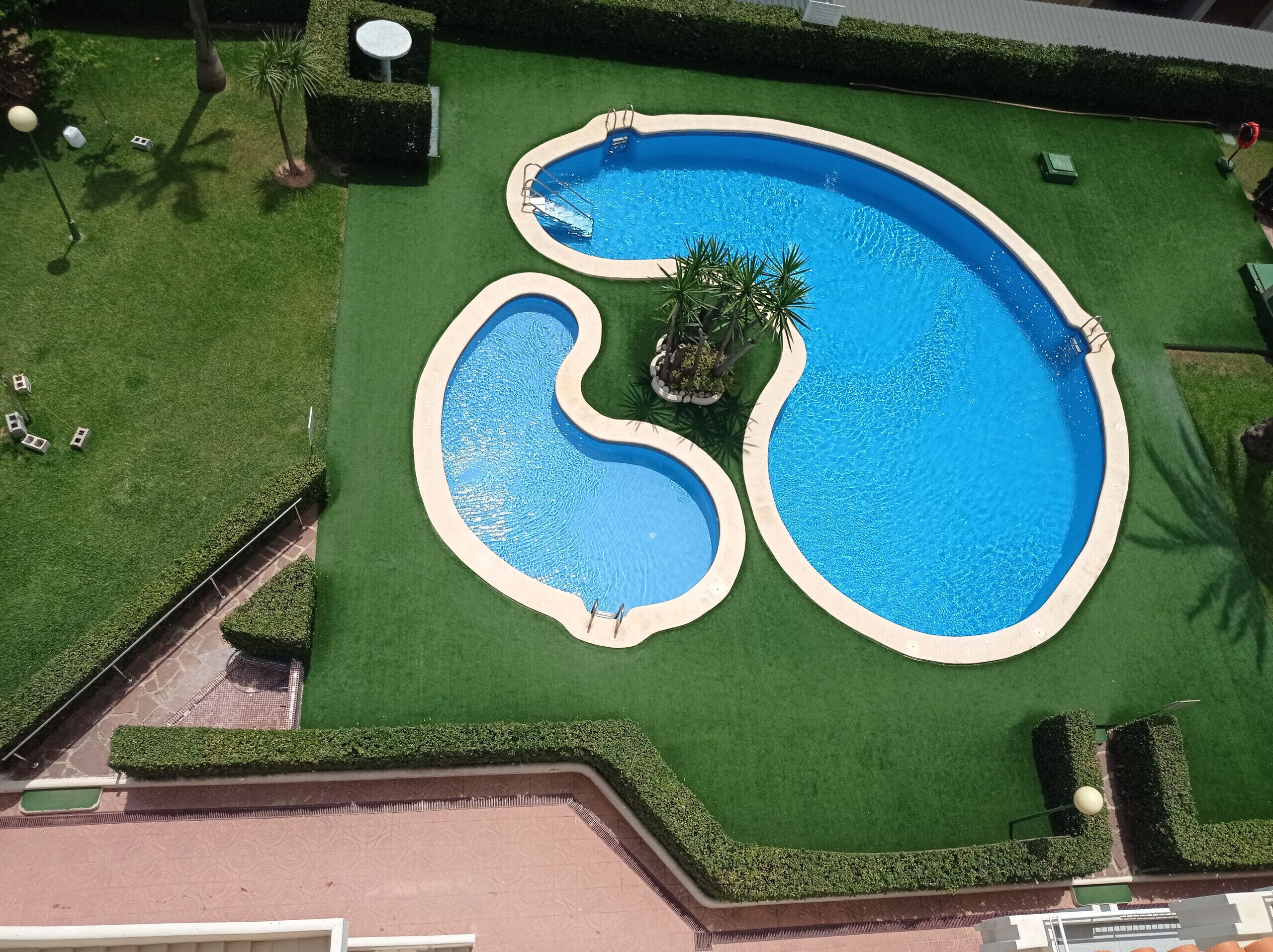 Une vue aérienne d’une piscine au design original, entourée d’une pelouse bien entretenue et de quelques arbres.