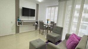 Un salon moderne doté d’un téléviseur à écran plat fixé au mur, d’un coin repas avec une table et des chaises, et d’une grande fenêtre laissant entrer la lumière naturelle.