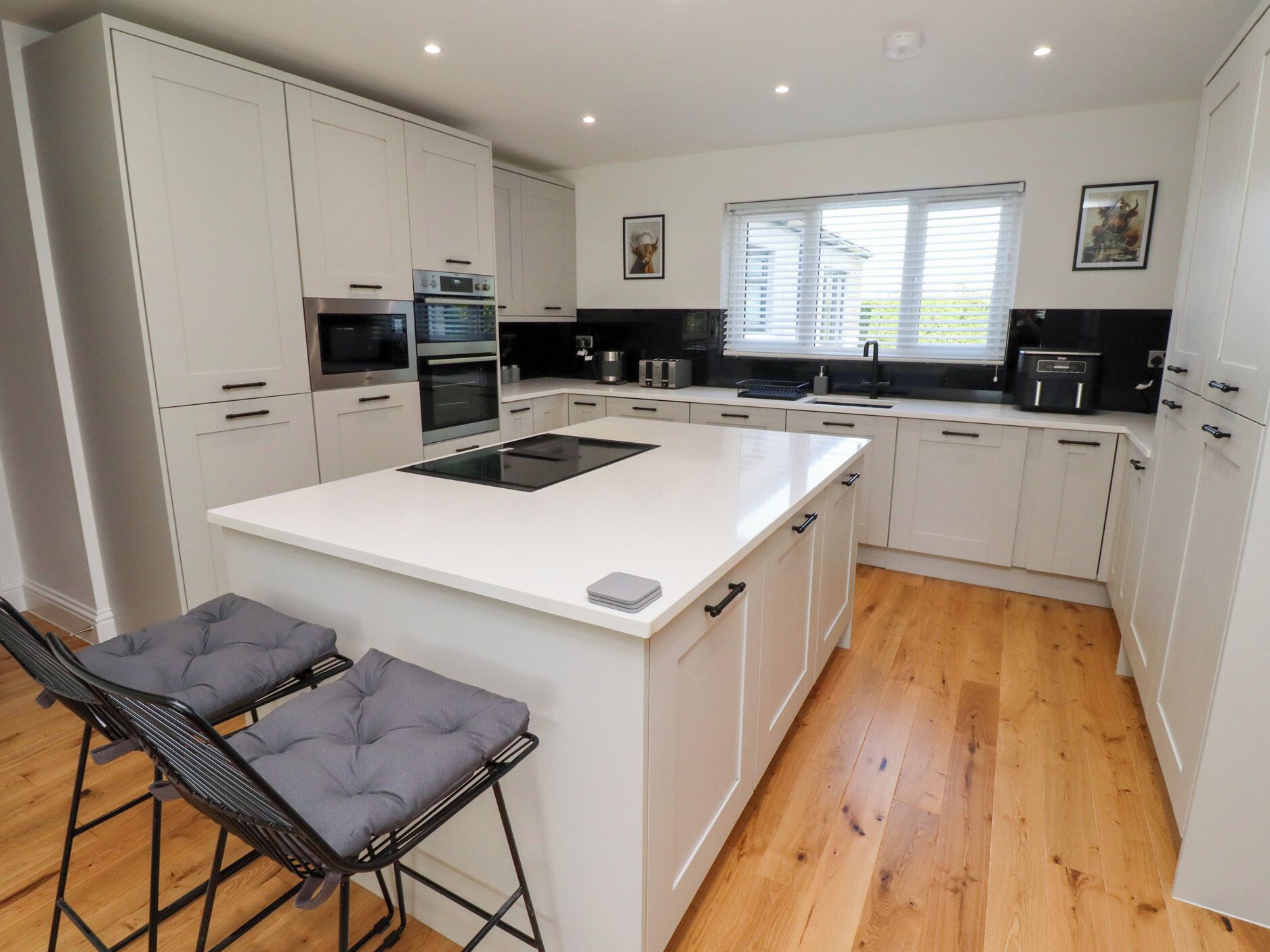A modern kitchen with white cabinetry, a central island, and a built-in oven. There are two bar stools with cushioned seats.