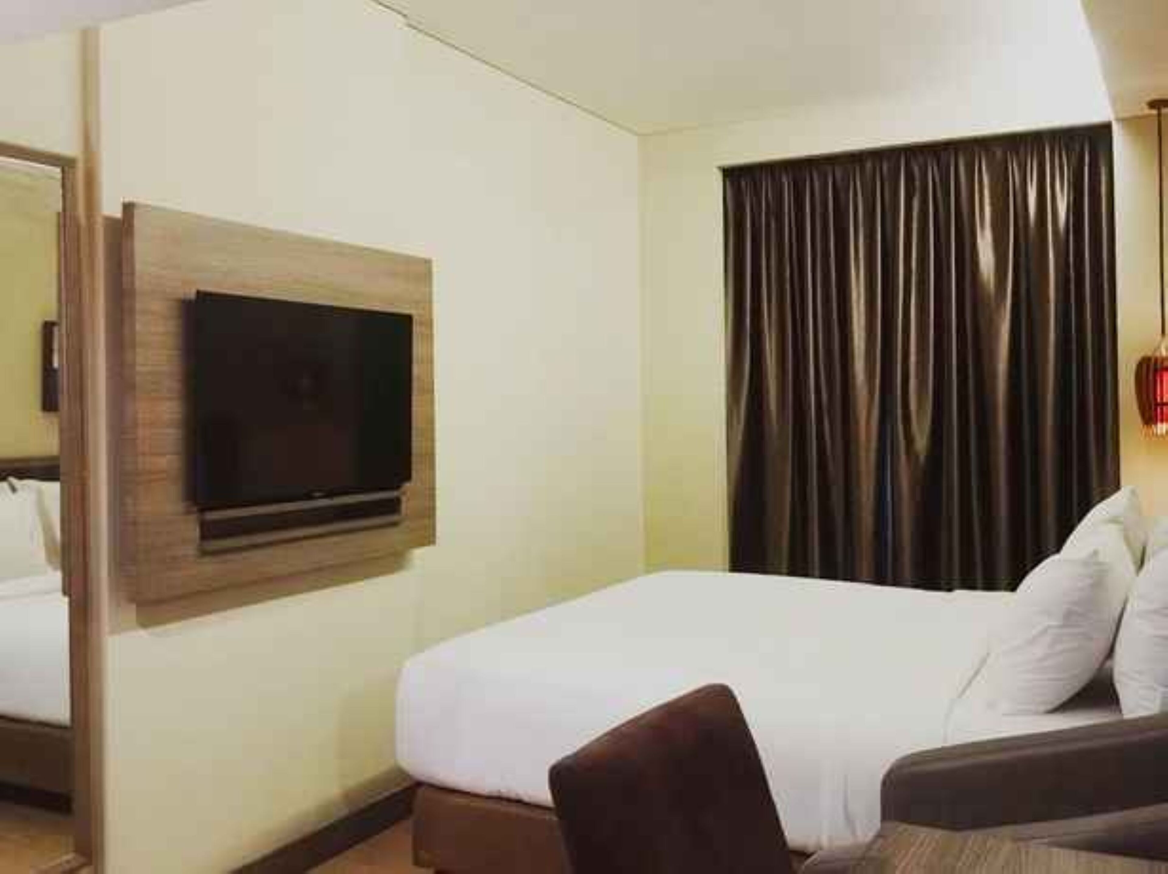 A hotel room with a bed, a flat-screen TV mounted on the wall, a large window with dark curtains, and a small table with a chair.