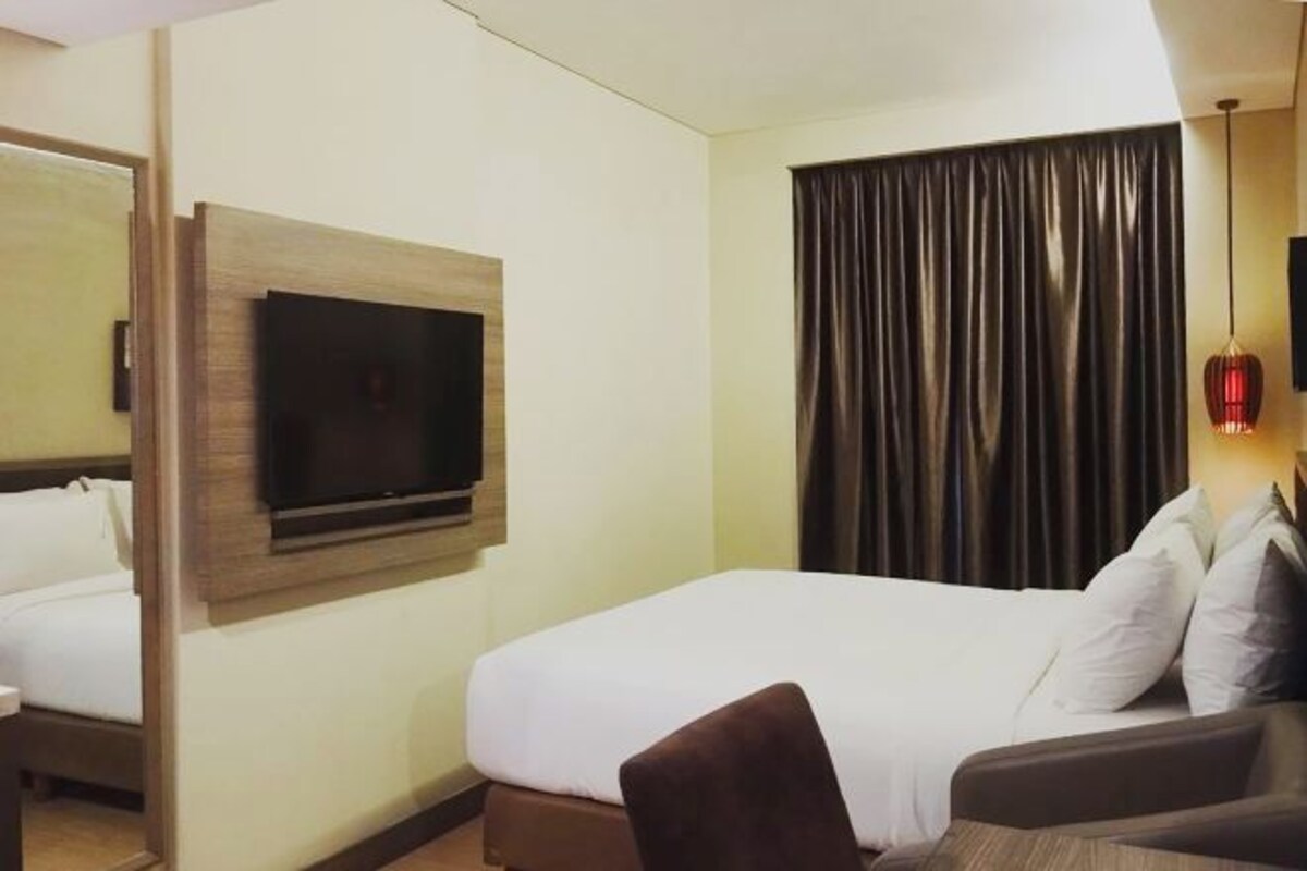 Hotellihuone, jossa on sänky, seinälle asennettu televisio, ikkuna verhoilla sekä pieni pöytä ja tuoli.