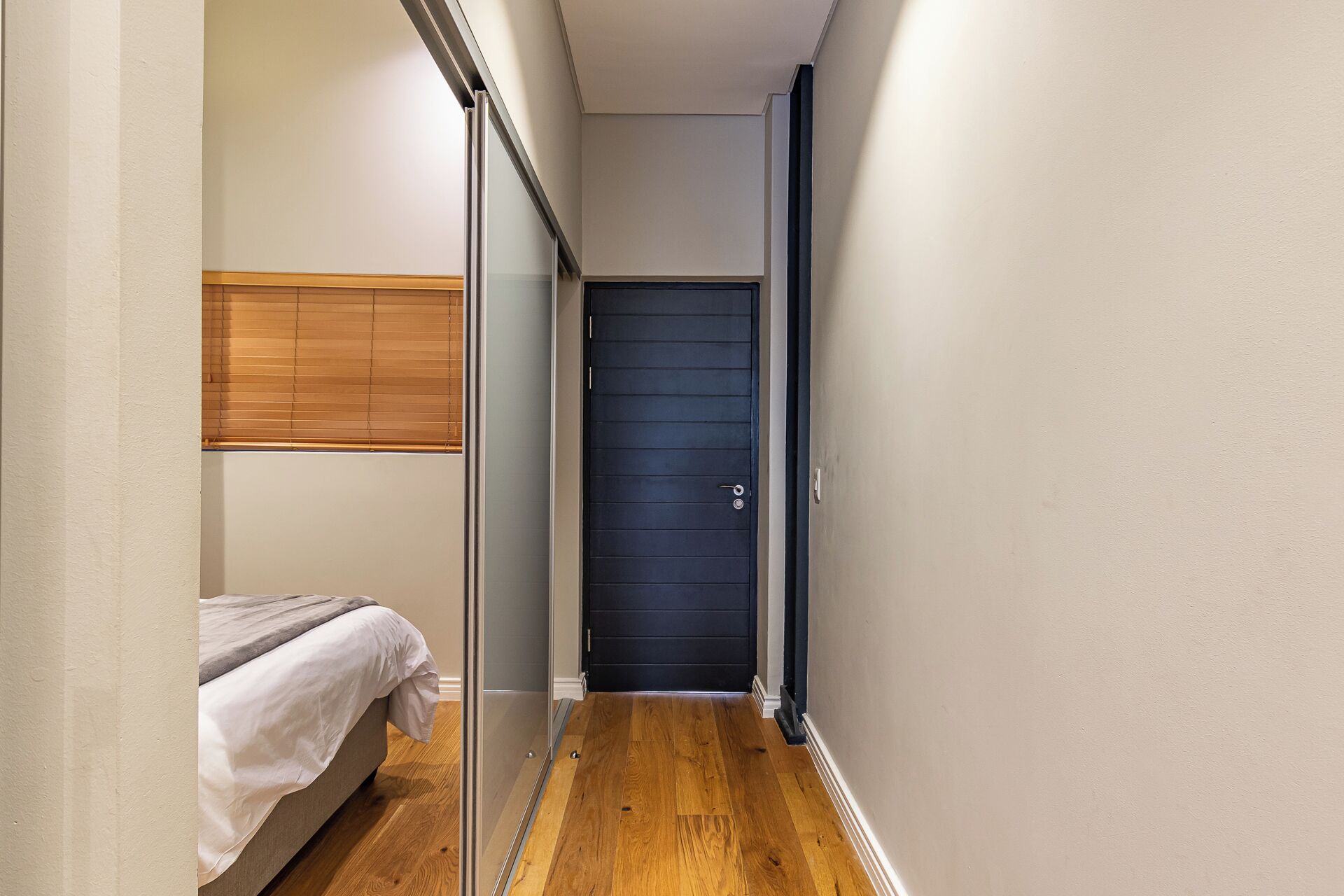 A narrow hallway with a bed on the left, a dark door at the end, and wooden flooring.
