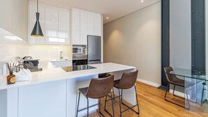A modern kitchen with white cabinets, a stainless steel refrigerator, and a glass dining table with two chairs. - The Docklands Luxury Apartments (Cape Town)