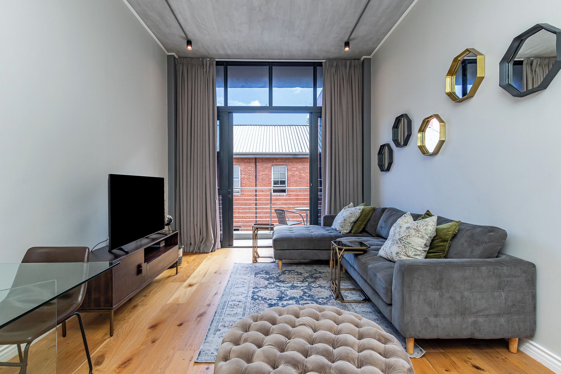 A modern living room with a grey sofa, a glass coffee table, a flat-screen TV, and a large window with curtains.