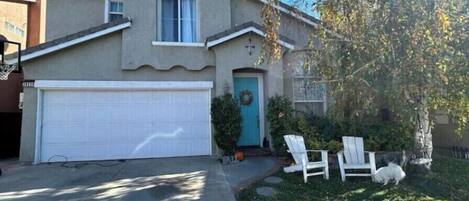 A two-story house with a garage, a front door with a wreath, and a small lawn with two white chairs and a dog.