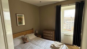 Een hotelkamer met een bed, nachtkastje, dressoir, raam met gordijnen en een foto aan de wand.