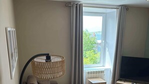 Een moderne woonkamer met een hoekbank, een televisie op een houten dressoir, een bijzettafeltje en een groot raam met gordijnen.